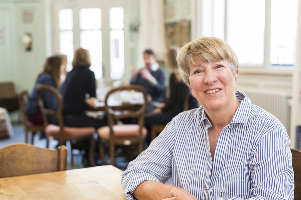 Ursula ist eine frisch gebackene Pensionärin aus Appenzell. Sie sitzt am Tisch am Erzählcafé, im Hintergrund ist die Gruppe in ein Gespräch vertieft. 